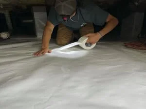Worker installing white polyethylene vapor barrier in crawl space
