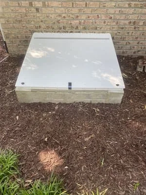 White plastic crawl space access cover with fasteners installed at ground level against brick foundation