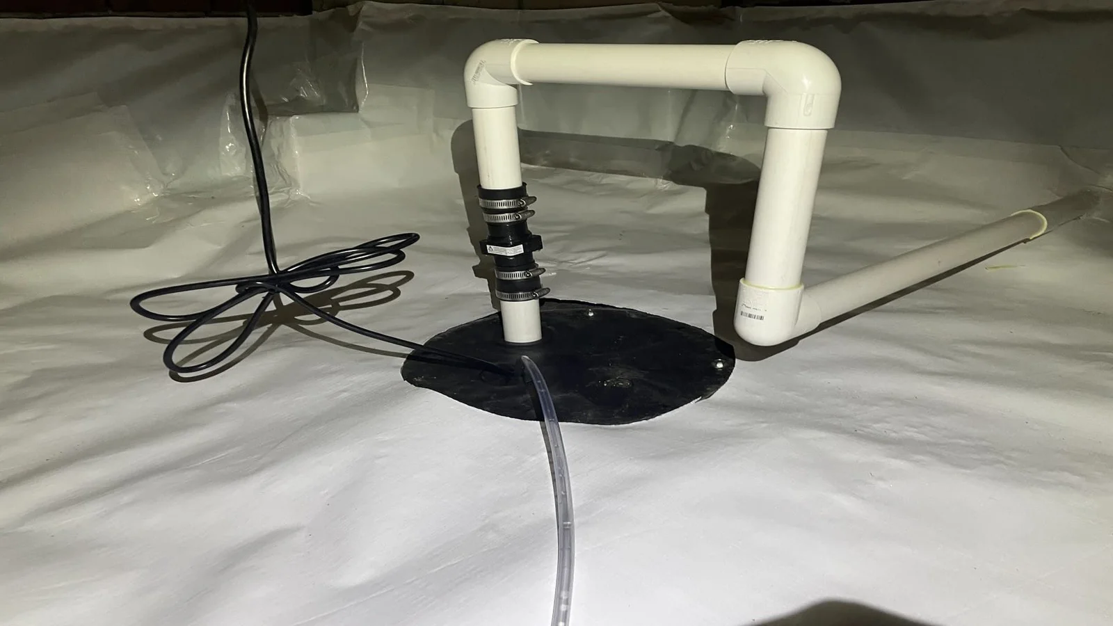 A sump pump installed in the basin of a crawl space beneath a Grimesland, NC home, with a discharge pipe routed toward the exterior foundation wall. The crawl space floor is clear of standing water.
